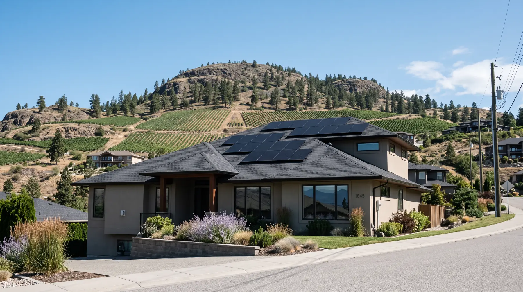 Black solar panels on a two-storey stucco home with vineyard slopes behind in West Kelowna